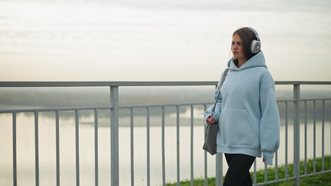 Young girl in blue hoodie with headphones walking beside river with iron rail demarcating her and water, scenic peaceful outdoor setting with flowing river and serene background view