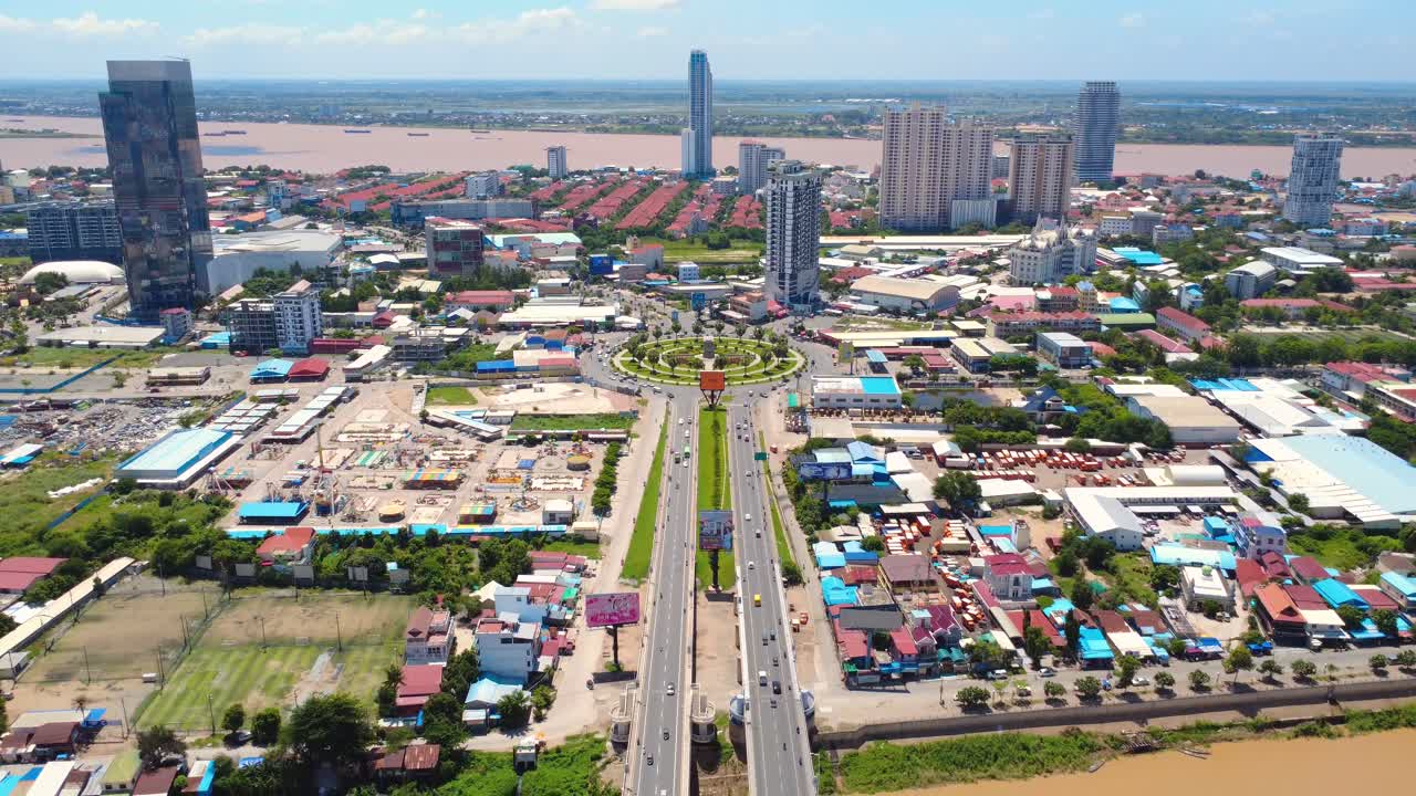 Drone video capturing vibrant Chroy Changvar cityscape with bustling roundabout, modern skyscrapers, and serene riverside under clear blue skies in Phnom Penh.