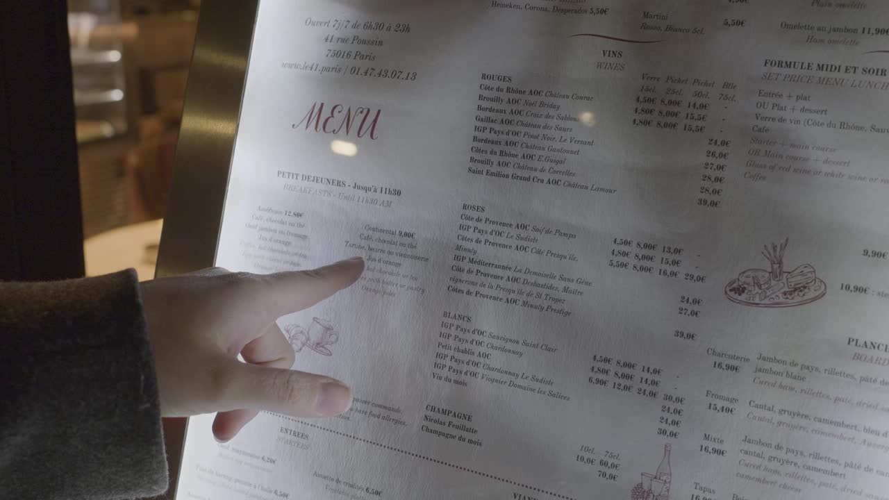 A hand pointing at a menu outside a café in Paris showcasing food options and beverages