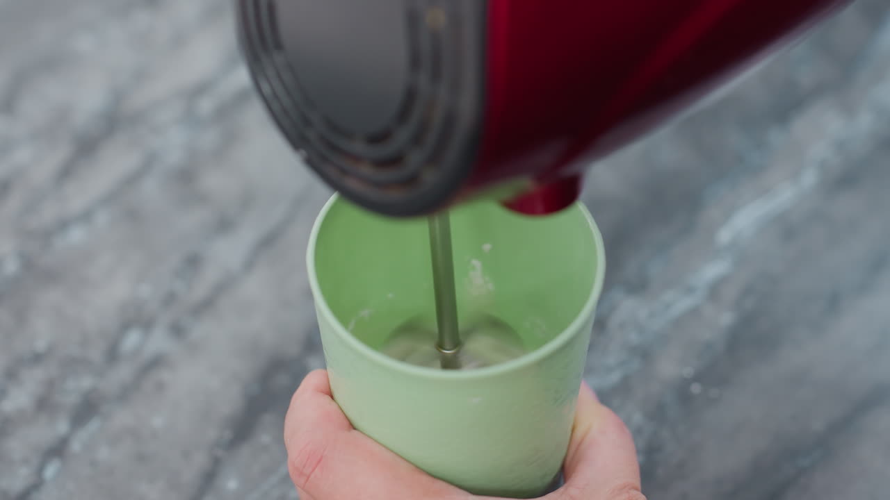 primer plano de una persona que utiliza un mezclador de mano eléctrico dentro de una taza verde, preparando bebidas o mezclando ingredientes, creando espuma, configuración de cocina, encimera con fondo borroso