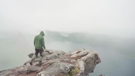 man hiker in hood walking towards the cliff ledge looking down the mountain on a foggy day at segla, senja, norway