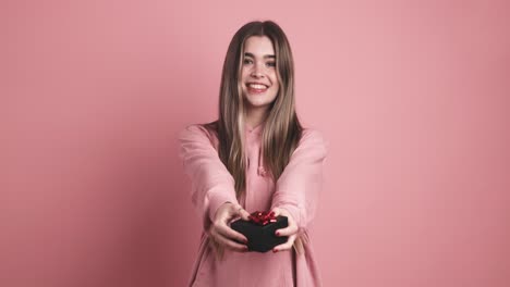 cheerful young woman giving gift box in pink studio