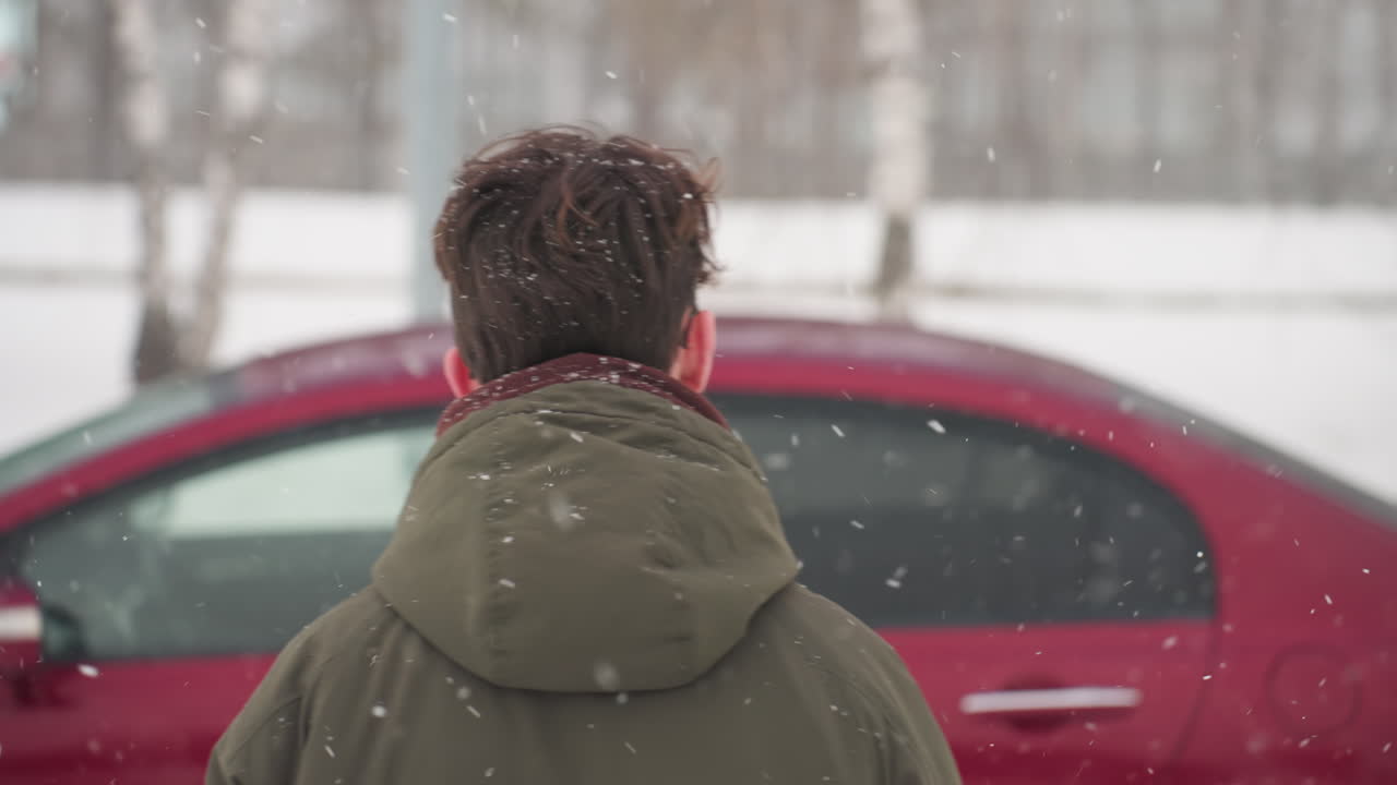 Rear view of boy wearing winter jacket walking across snowy outdoor parking lot toward parked car during snowfall with blurred trees in background capturing cold winter journey outdoors
