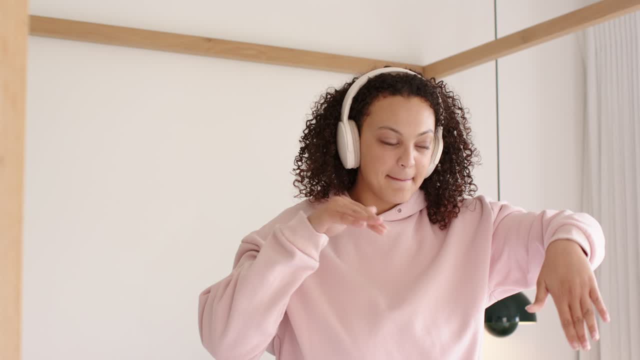 Donna afroamericana con felpa rosa con cappuccio che indossa cuffie bianche e si muove a ritmo di musica in camera da letto
