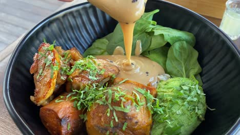 peanut sate sauce being poured over healthy food bowl with brown rice, chicken, sweet potato, spinach, and avocado, in classy black bowl