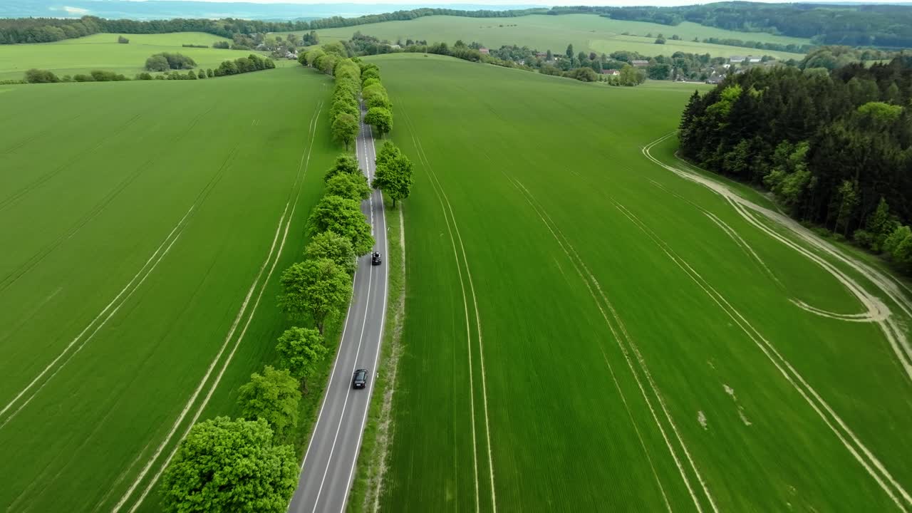 Cars driving along a country road lined with trees, surrounded by green fields and rural landscape, seen from a bird’s-eye drone perspective.