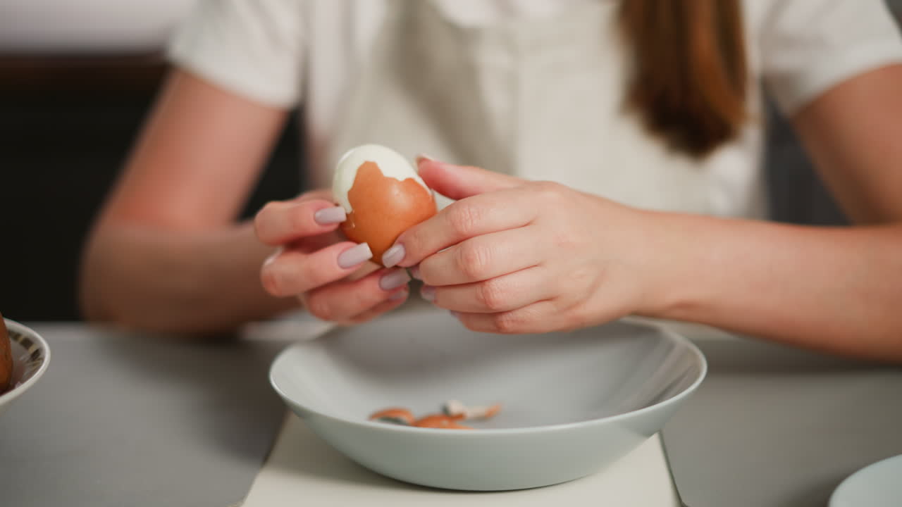 Close up of fair skinned woman peeling boiled egg over dish with egg shell pieces inside, wearing white shirt and apron, blurred view of brown hair flowing down and edge of another plate