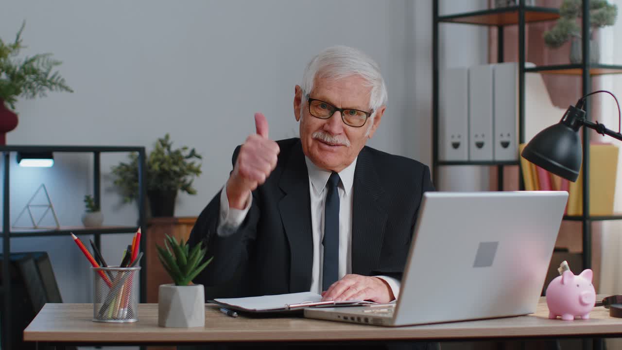 Senior business man raises thumbs up agrees approve likes good news using laptop at home office