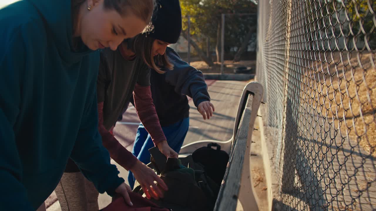 A trio of girls in sports uniforms put their sports bags on a bench near a lattice fence on a summer outdoor sports ground and prepare for basketball practice in the afternoon