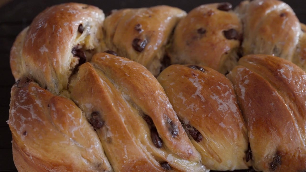 Primo piano estremo con carrellata sui fili intrecciati lucidi di una brioche con gocce di cioccolato che si raffredda su una griglia metallica