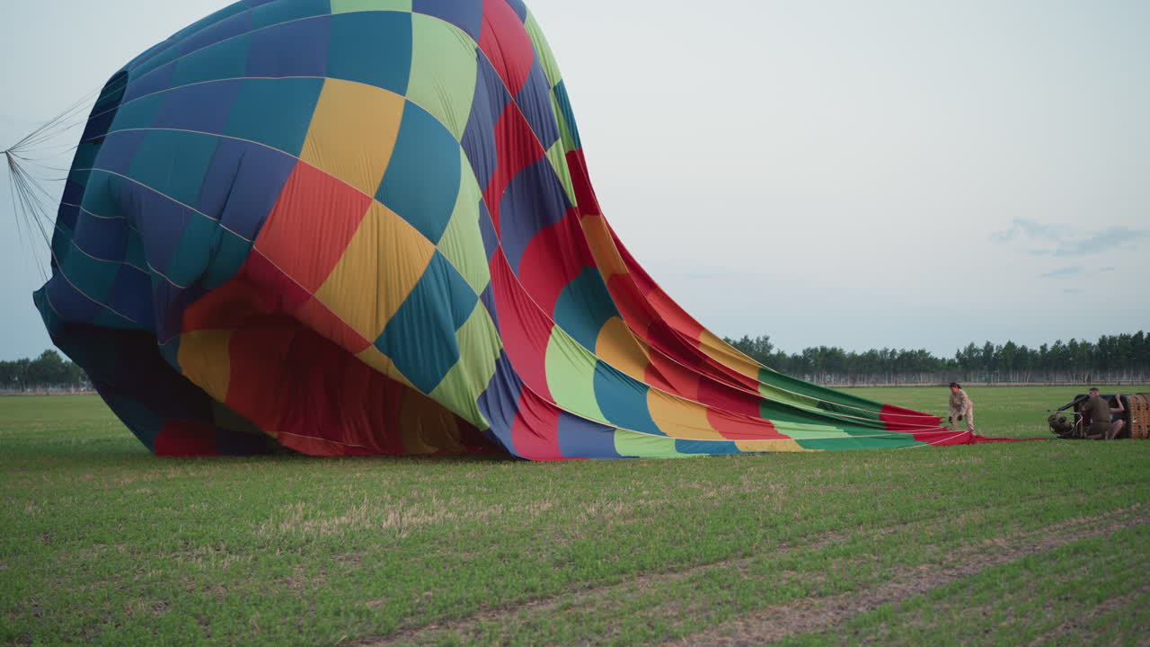 woman pulling tether while deflating vibrant hot air balloon canopy onto green field with male assistant adjusting basket and preventing envelope drift during ground