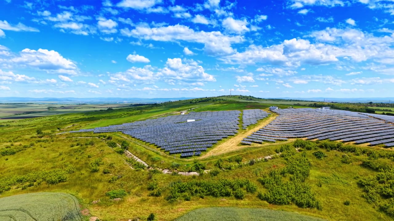 Alternativ solenergipark på grøn bakkeskråning med skyer. Vidvinkel luftfoto af rækker af solpaneler på grøn græsklædt bakke under blå himmel.