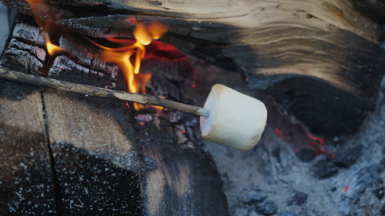 Close up of a marshmallow roasting on a stick near open wood fire in slow motion.