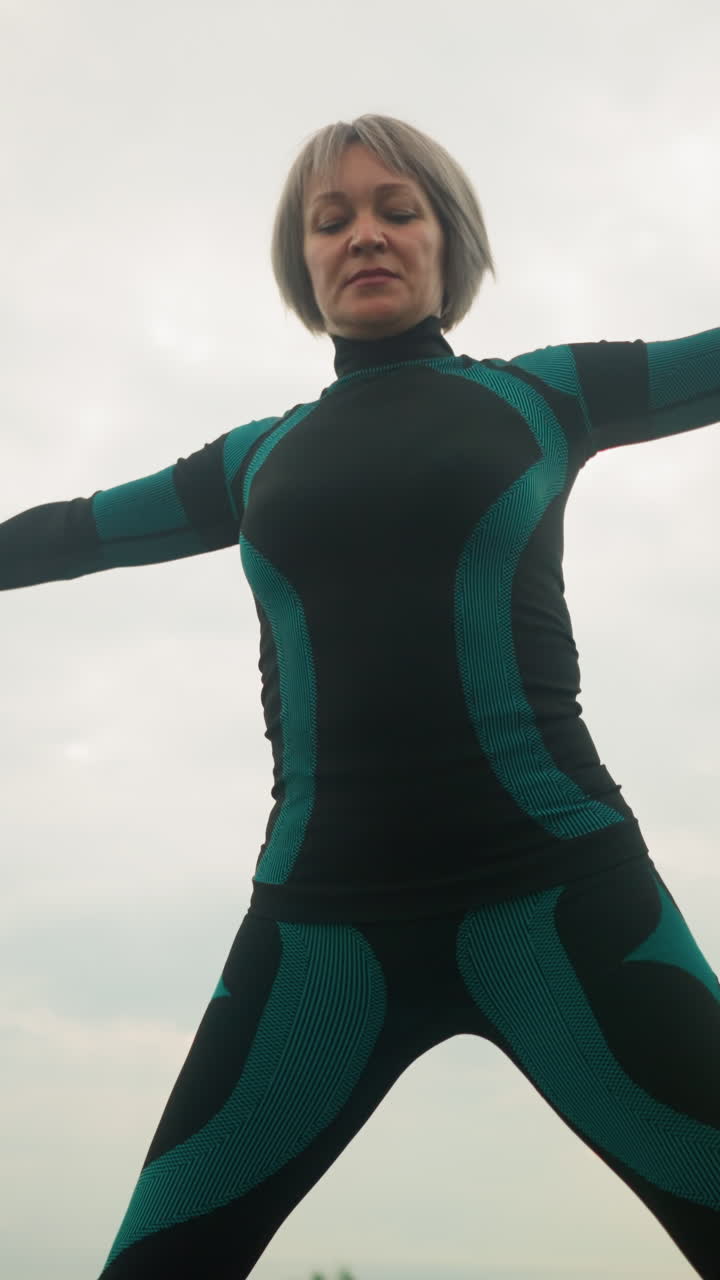 Low-angle view of a woman in a green-black suit standing on a yoga mat, practicing yoga triangle pose with arms open wide and legs apart, set against a cloudy sky