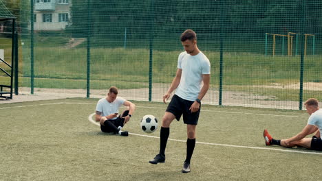 young street football player training freestyle tricks while two team players sitting on the pitch doing stretch exercises and another one using mobile phone