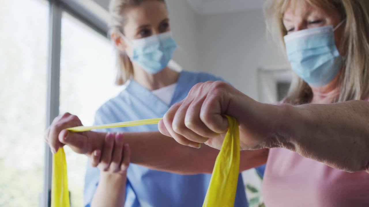 여성 의료 종사자가 고령 여성에게 집에서 운동 밴드를 사용하여 운동을 도와줍니다.