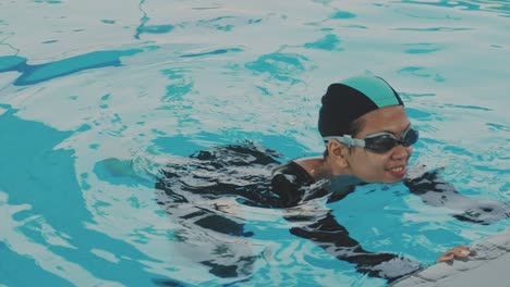 Una-Joven-Asiática-Usa-Gafas-Y-Gorra,-Aprende-A-Nadar-Sosteniendo-Junto-A-La-Piscina-Y-Mueve-Las-Piernas-Bajo-El-Agua