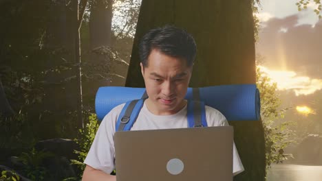 close up of asian male hiker with mountaineering backpack using a laptop while exploring forest nature