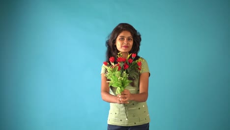 A-young-Indian-girl-in-green-t-shirt-standing-with-a-bouquet-of-flowers-smiling-at-the-camera-in-an-isolated-blue-background-studio