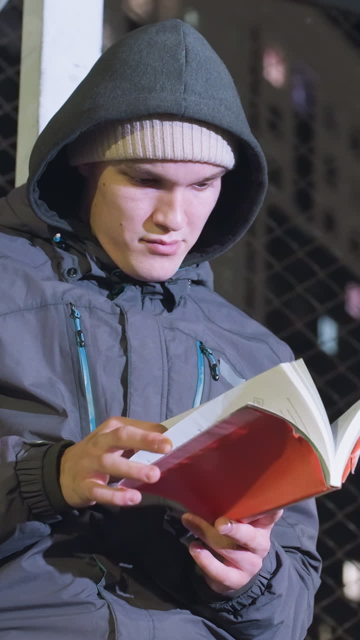hombre con capucha y gorra apoyado en un poste de meta metálico, leyendo un libro contemplativamente al aire libre por la noche, revisando las páginas bajo las luces de la ciudad urbana con un fondo de valla de malla