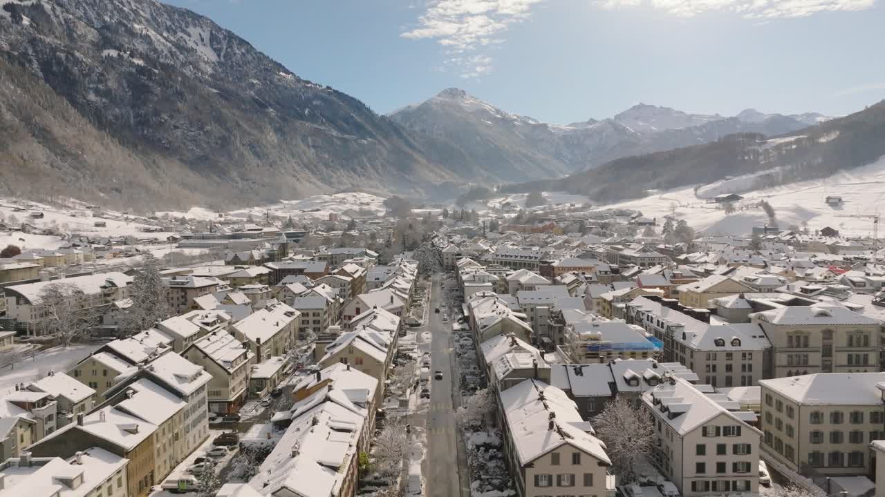 Aerial footage of the snow-covered town of Glarus in the Swiss Alps. The drone moves slowly forward on a clear winter day, showing rooftops, streets, and surrounding mountain scenery