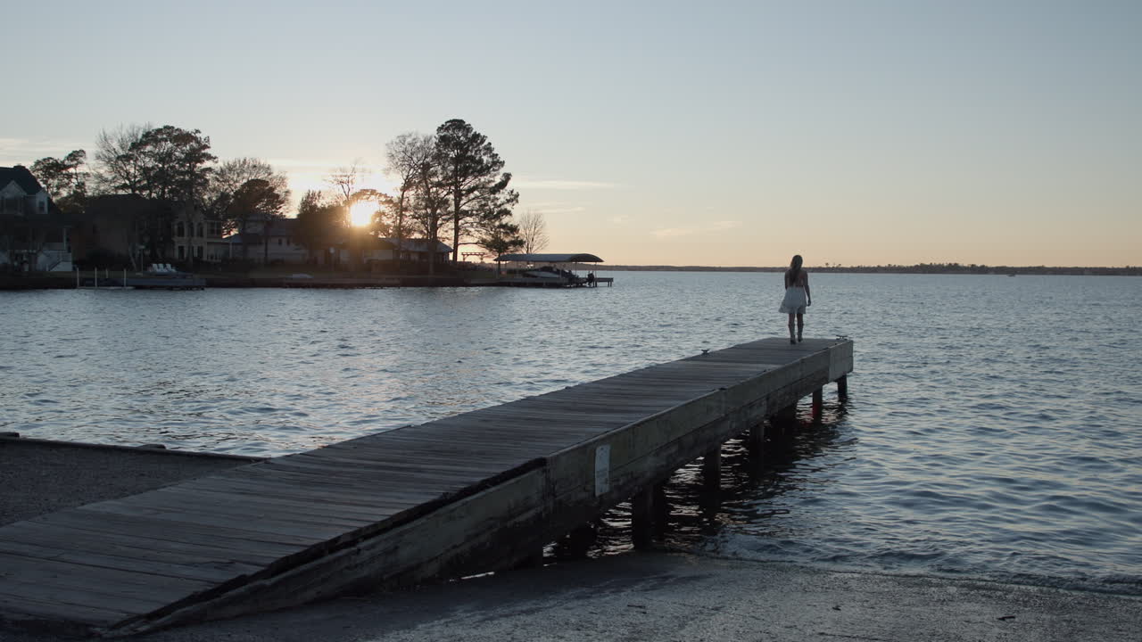 Ampia inquadratura di una bella ragazza bionda che cammina lungo un pontile per barche al tramonto.