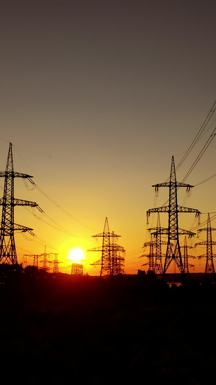 Electric pylons against orange sunset. Dark background of high-voltage electric lines in the evening. Transmission lines on the setting sun. Vertical video
