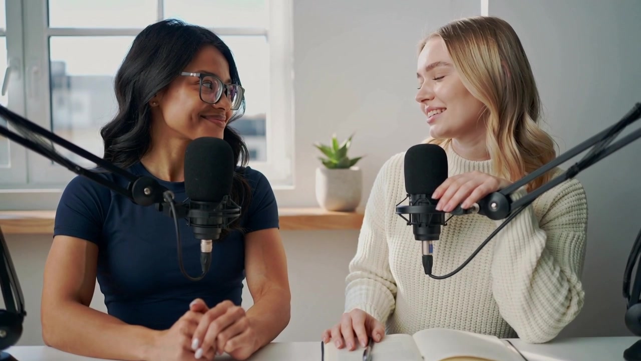 Verten til høyre i genser ser på notatbok og leser inn i mikrofon, mens en kollega ser på i et studio for podcast.