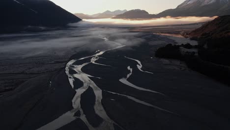 Aerial-view-of-the-braided-Bealey-river-in-Arthurs-pass-at-sunrise-in-a-valley-surrounded-by-mountains,-flying-towards-the-mountains-and-the-sun