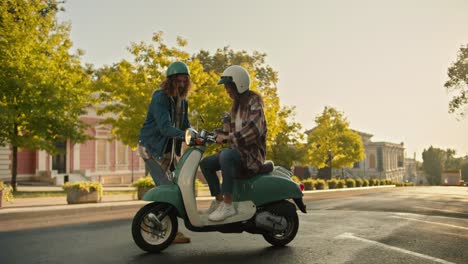 A-guy-with-curly-hair-in-denim-helps-a-girl-move-while-riding-a-moped-on-the-street-in-summer.-A-guy-in-a-checkered-shirt-helps-his-girlfriend-ride-a-moped-and-teaches-her-on-a-wide-street-with-trees-at-sunrise-in-summer