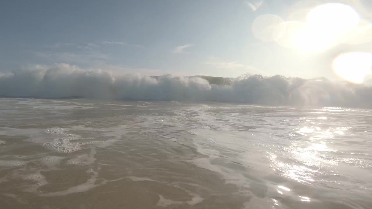 A backlit shorebreak swell advances toward a low waterline camera at Mirissa Beach, Sri Lanka, growing into a foamy wall beneath bright tropical sun and ocean glare.
