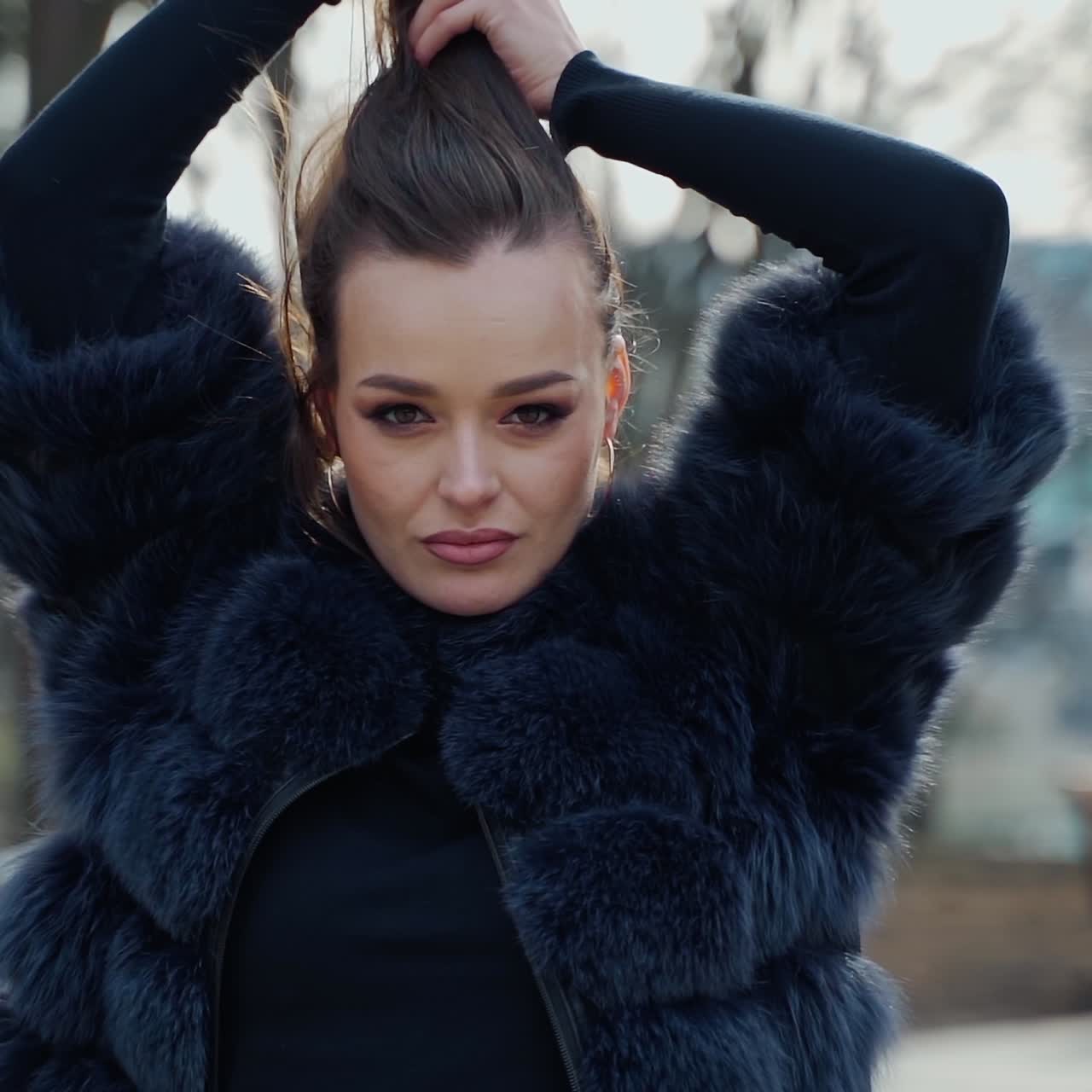 Portrait of a woman outdoors. Sexy girl in black fur coat looking on camera while holding her brown hair on the city park background. Slow motion.