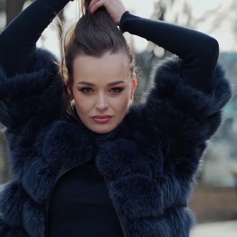 portrait of a woman outdoors. sexy girl in black fur coat looking on camera while holding her brown hair on the city park background. slow motion.