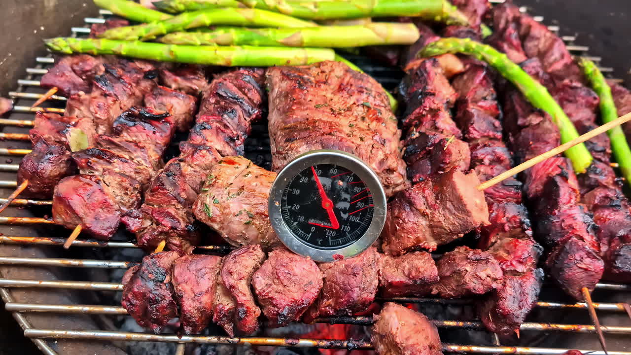 Close-up of delicious beef shish kebabs and fresh green asparagus cooking on a hot charcoal grill, with a thermometer checking the meat's internal temperature for perfect doneness