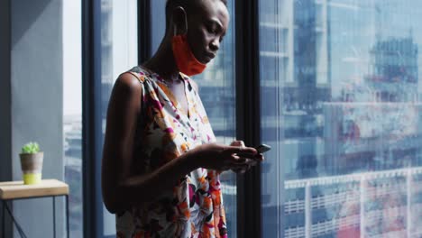 African-american-woman-with-face-mask-around-her-neck-using-smartphone-at-modern-office