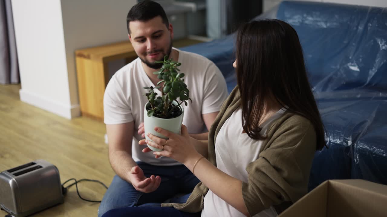pareja alegre sentada en el suelo de la sala de estar, desempaquetando la planta de la caja
