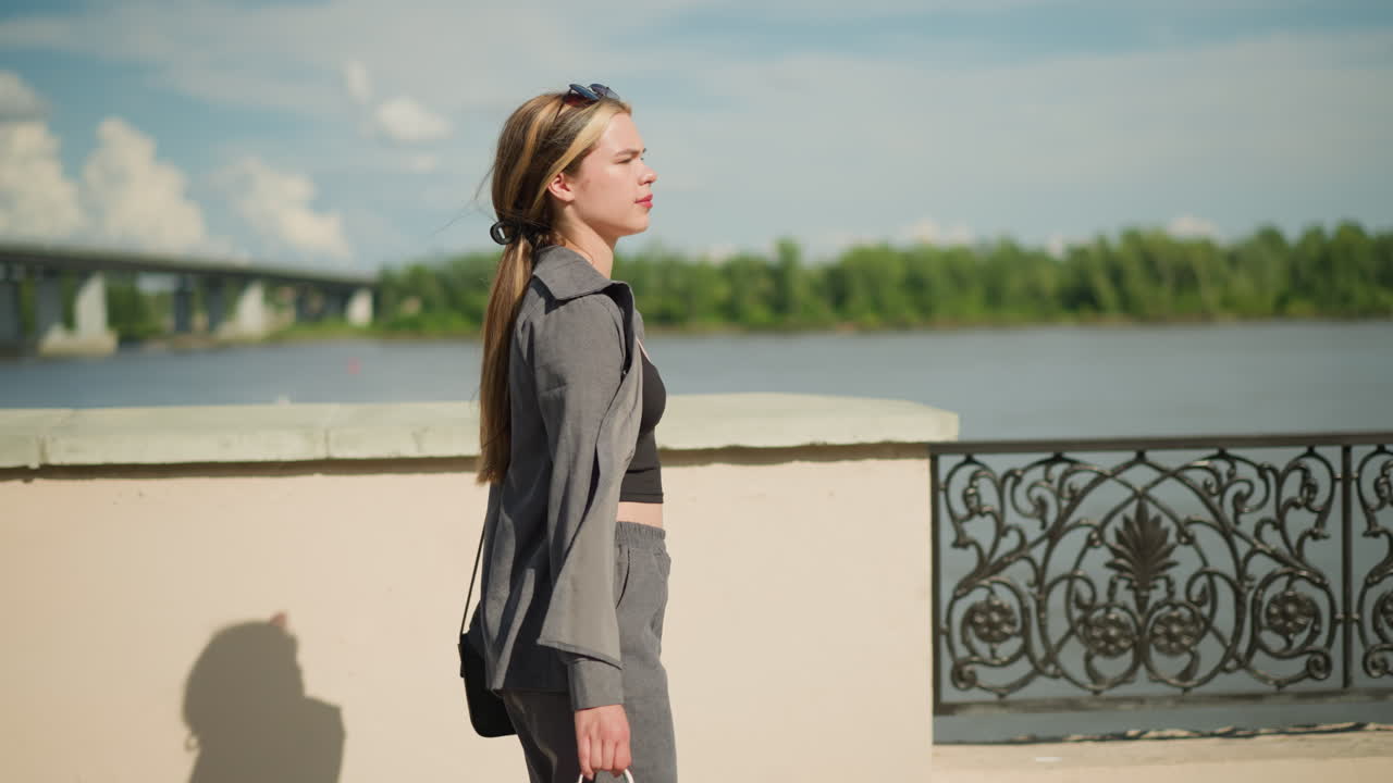 Lady walking near riverfront with shopping bag in hand, passing a decorative metal fence, background features blurred bridge, greenery, and clear sky on a sunny day, while she gazes towards the river