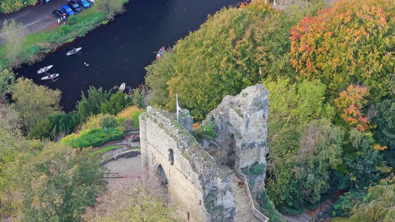 High angle drone shot shows aged stone walls and partial towers of Knaresborough Castle, complete with flag waving over the ruins, along with mature riverside trees and boats on the River Nidd