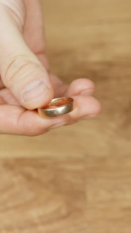 a hand gently holds a polished golden ring against a wooden table background. captured in vertical format, the frame emphasizes simplicity and detail in an intimate, minimalistic setting.