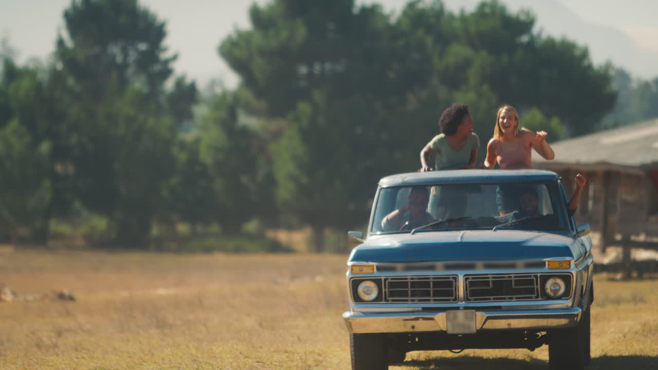 Zwei Frauen stehen auf der Ladefläche eines Pick-up-Trucks, während Freunde einen Roadtrip zu einer Landhütte genießen