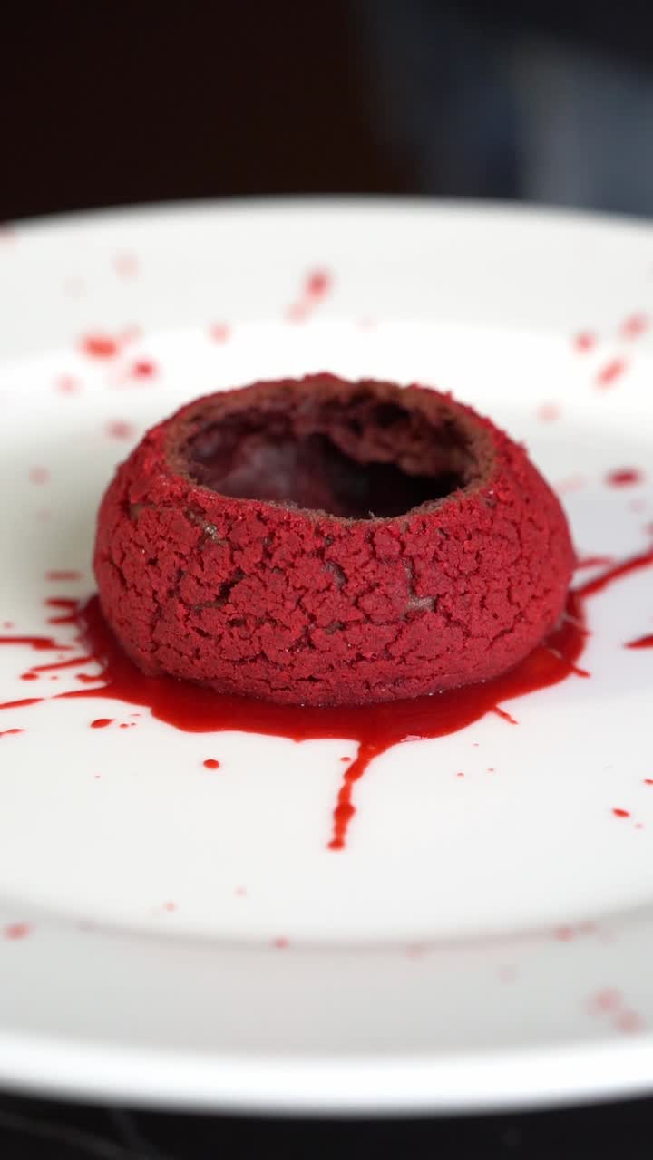 Un chef prepara un postre rojo en un plato blanco, añadiendo relleno dentro de un pastel de terciopelo rojo con salsa salpicada alrededor, primer plano. Insertar toma.