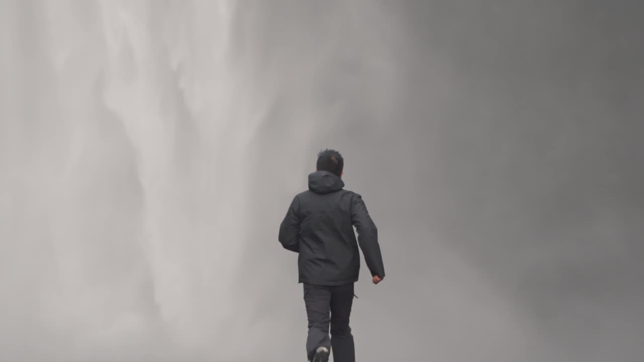 Man in outdoor gear, seen from behind, running purposefully towards the powerful mist of Skógafoss waterfall in Iceland