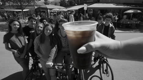 hand holding a colored drink in front of a monochrome group of young friends with bicycles