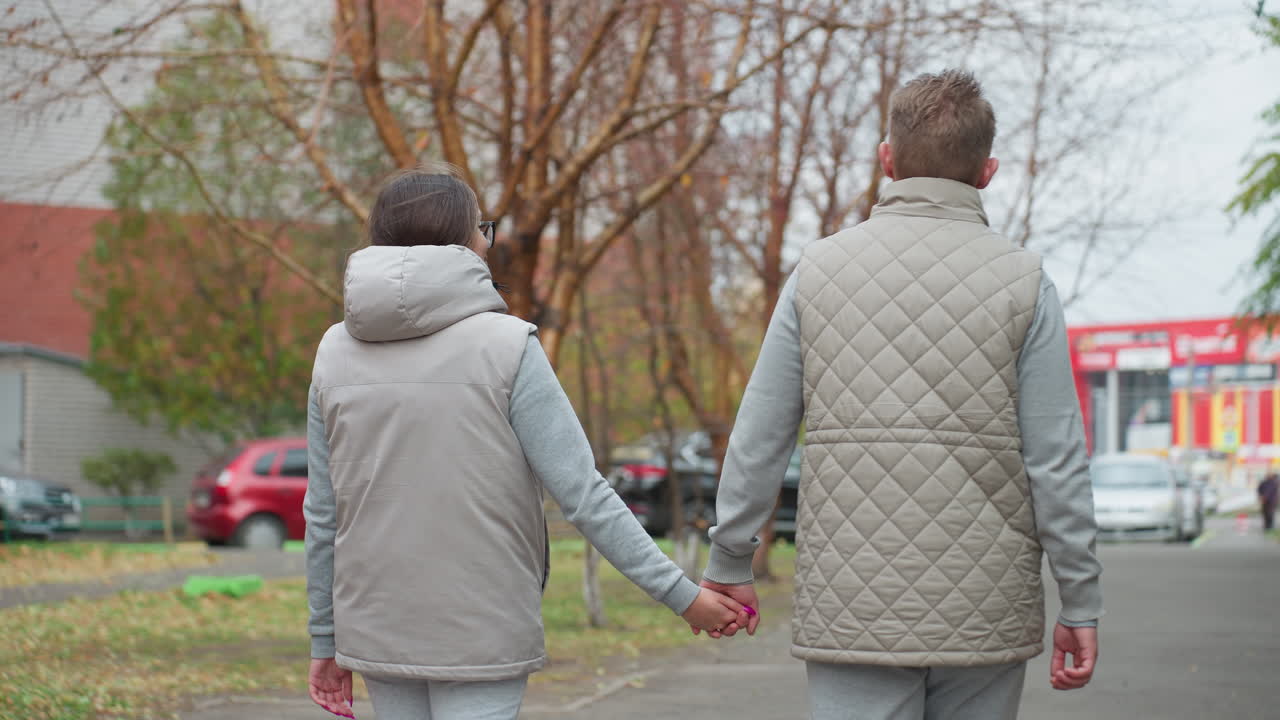 Back view of two couples walking hand in hand along quiet street in light-colored matching outfits, surrounded by trees and parked cars with soft natural lighting