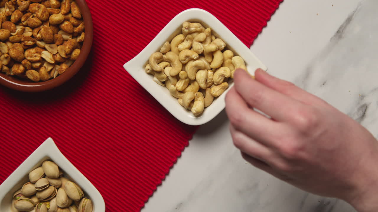 Close Up Of Hand Choosing From Bowls Of Cashews Dry Roasted Peanuts And Pistachio Nuts In Studio