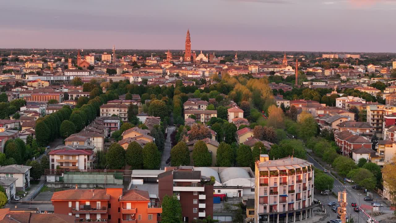 Golden sunlight illuminating historic rooftops and cobblestone streets of Cremona, Torrazzo tower rising above urban landscape during twilight, drone ascending shot