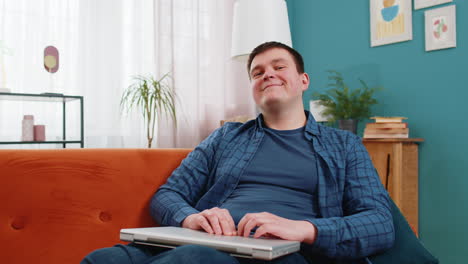 happy freelancer man sitting on sofa closing laptop and relaxing after finishing work job at home