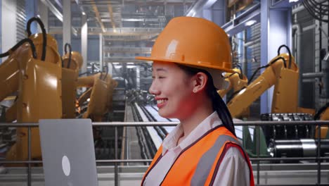 close up side view of asian female engineer with safety helmet working on a laptop and looking around while standing with industrial robotic arms working