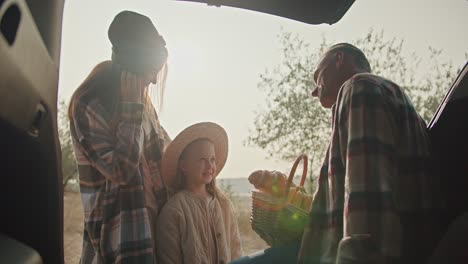 A-happy-family,-middle-aged-man-with-gray-hair-in-a-checkered-shirt-communicates-with-his-little-daughter,-a-blonde-girl,-and-with-his-wife,-a-girl-with-brown-hair-in-a-checkered-shirt.-Sitting-in-the-open-trunk-of-a-car-during-his-vacation-and-picnic-outside-the-city