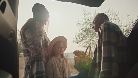 a happy family, middle-aged man with gray hair in a checkered shirt communicates with his little daughter, a blonde girl, and with his wife, a girl with brown hair in a checkered shirt. sitting in the open trunk of a car during his vacation and picnic outside the city